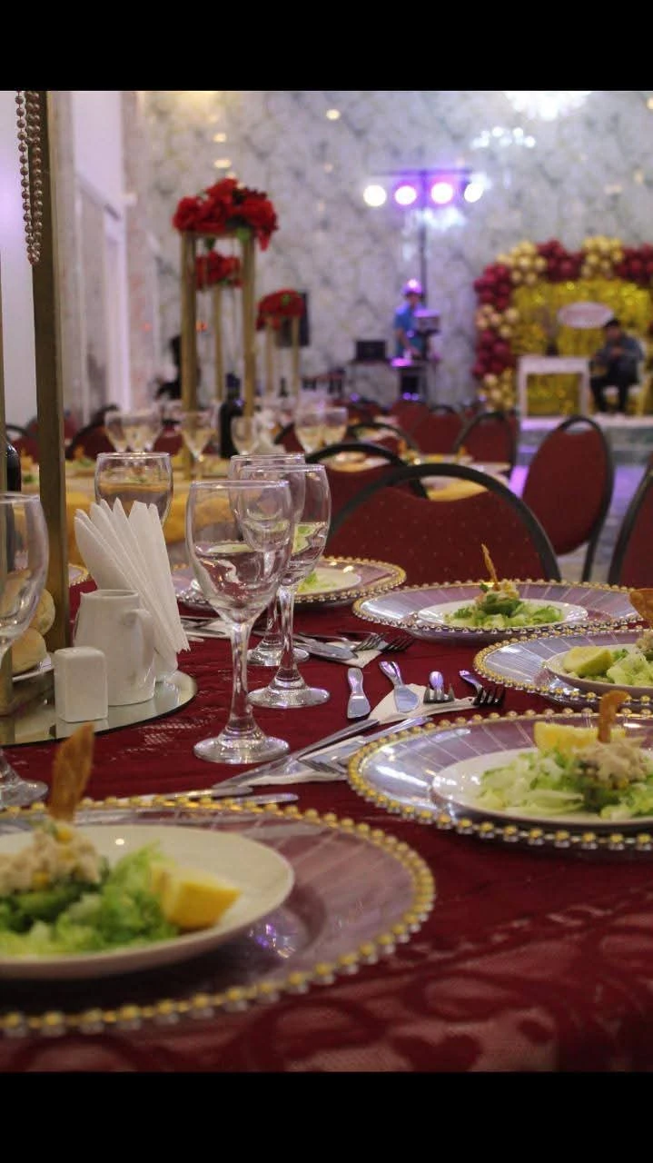 Mesa de cena corporativa con mantelería burdeos, copas y platos servidos; centros florales rojos y luces al fondo.