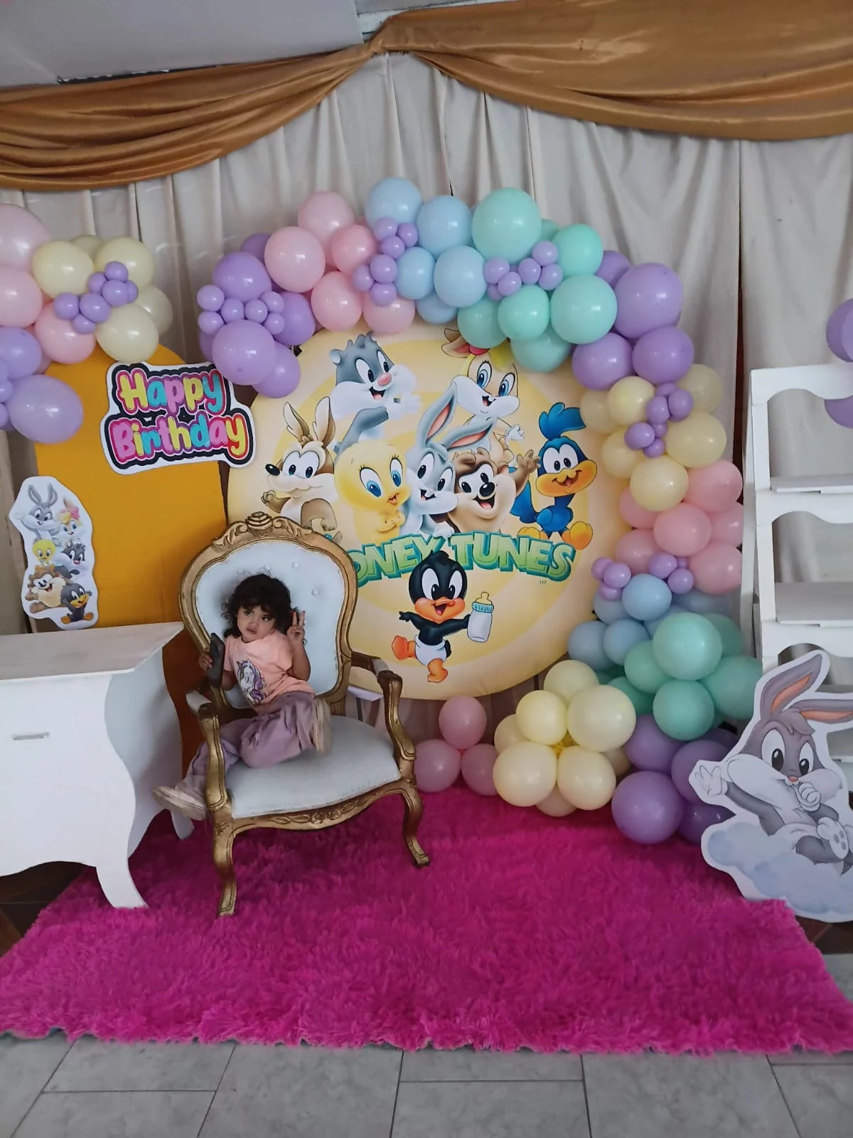 Montaje de cumpleaños con personajes de Baby Looney Tunes, globos pastel y una niña en una silla blanca con marco dorado.