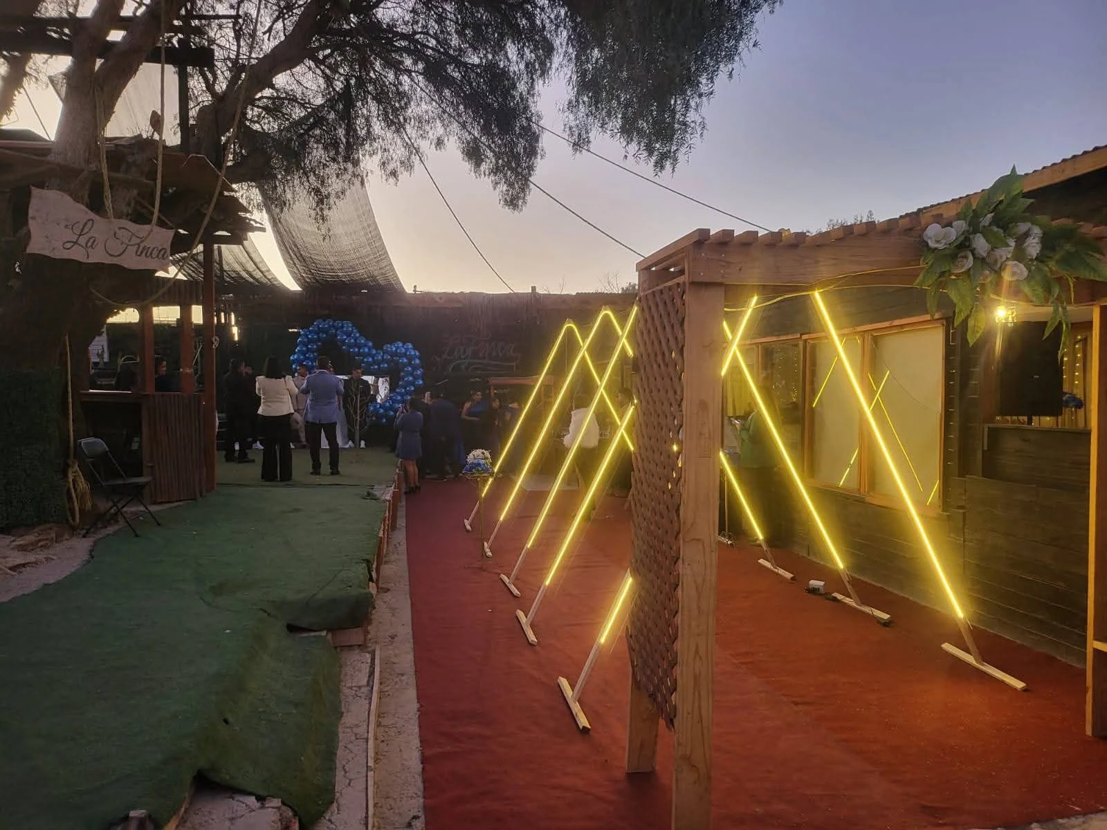 Espacio exterior de Manantial al atardecer, con construcciones de madera e iluminación amarilla en forma de rombos.