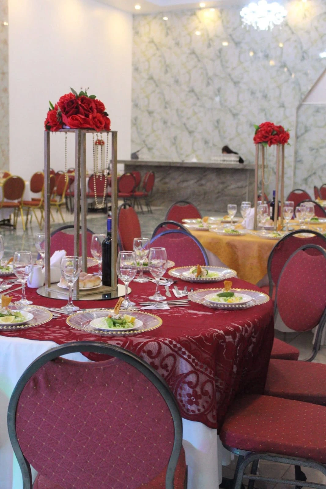 Mesa con mantelería burdeos, platos servidos, copas y centros florales rojos altos.