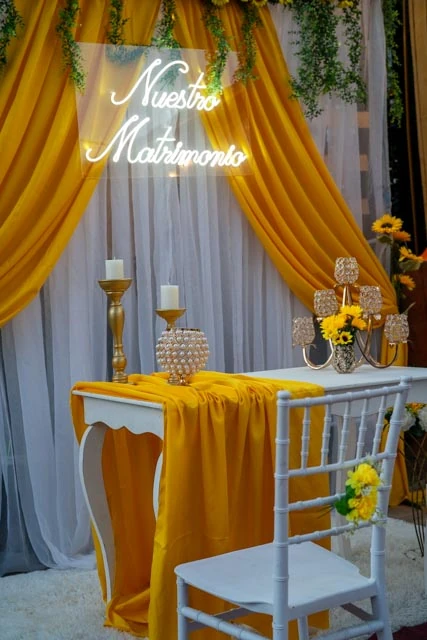 Mesa para novios con telas doradas y blancas, girasoles y un letrero luminoso de recién casados.